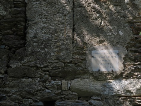 Lapide commemorativa su un muro di pietra, illuminata dalla luce, con dettagli architettonici rustici.