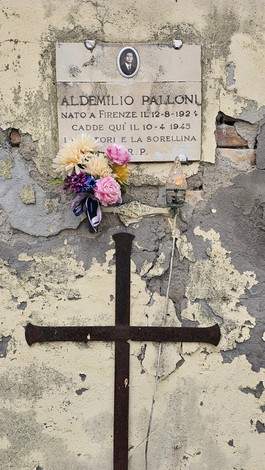 Croce in ferro con ai piedi fiori e una foto affissa su un muro degradato.