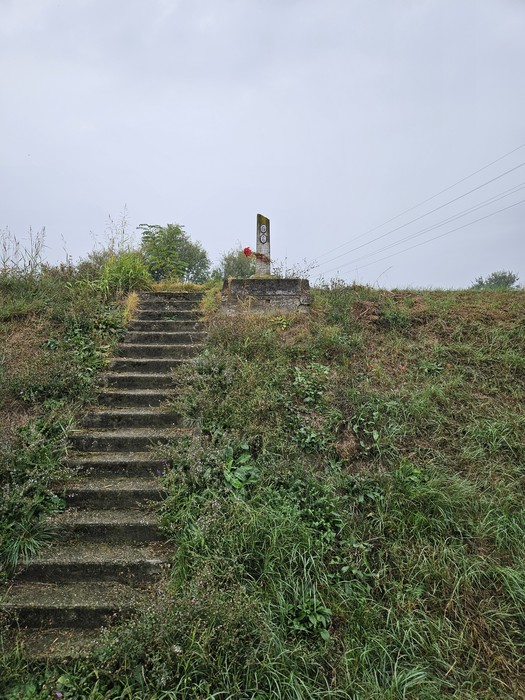 Scalinata in pietra tra l'erba porta a un cippo con iscrizione, in cima a un rilievo erboso.