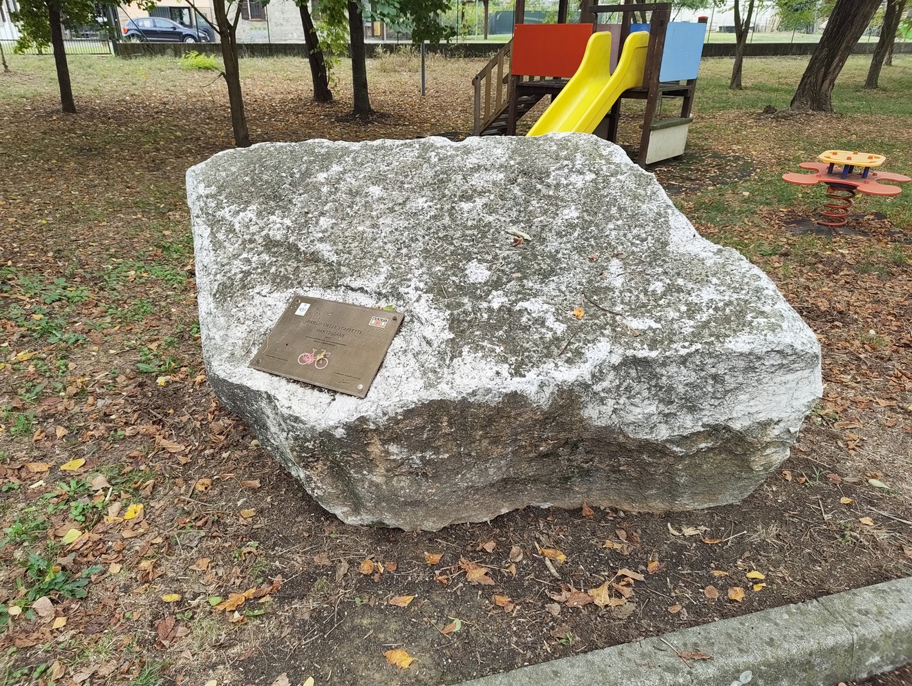 Grande masso di pietra con targa commemorativa, in un parco con giochi per bambini sullo sfondo.
