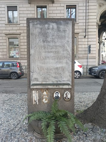 Stele commemorativa con nomi incisi, circondata da elementi naturali e architettura urbana sullo sfondo.