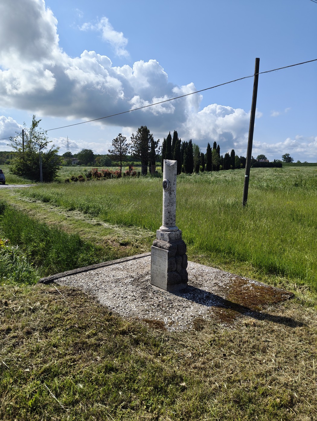 Cippo storico in un campo verde, circondato da alberi e nuvole bianche nel cielo.