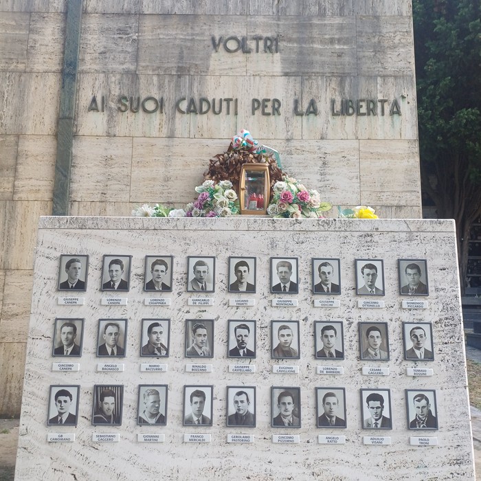 Sacrario in pietra con iscrizione, teca con fiori e pannello con foto e nomi dei caduti.