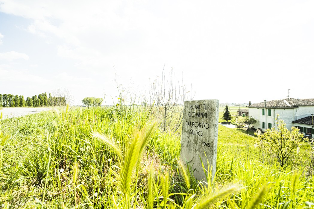 Cippo commemorativo a Romitti e Dalporto immerso nella natura, con case sullo sfondo.