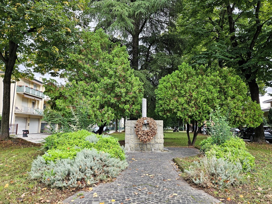 Cippo in pietra con corona in un'aiuola, tra alberi, al termine di un vialetto pedonale.