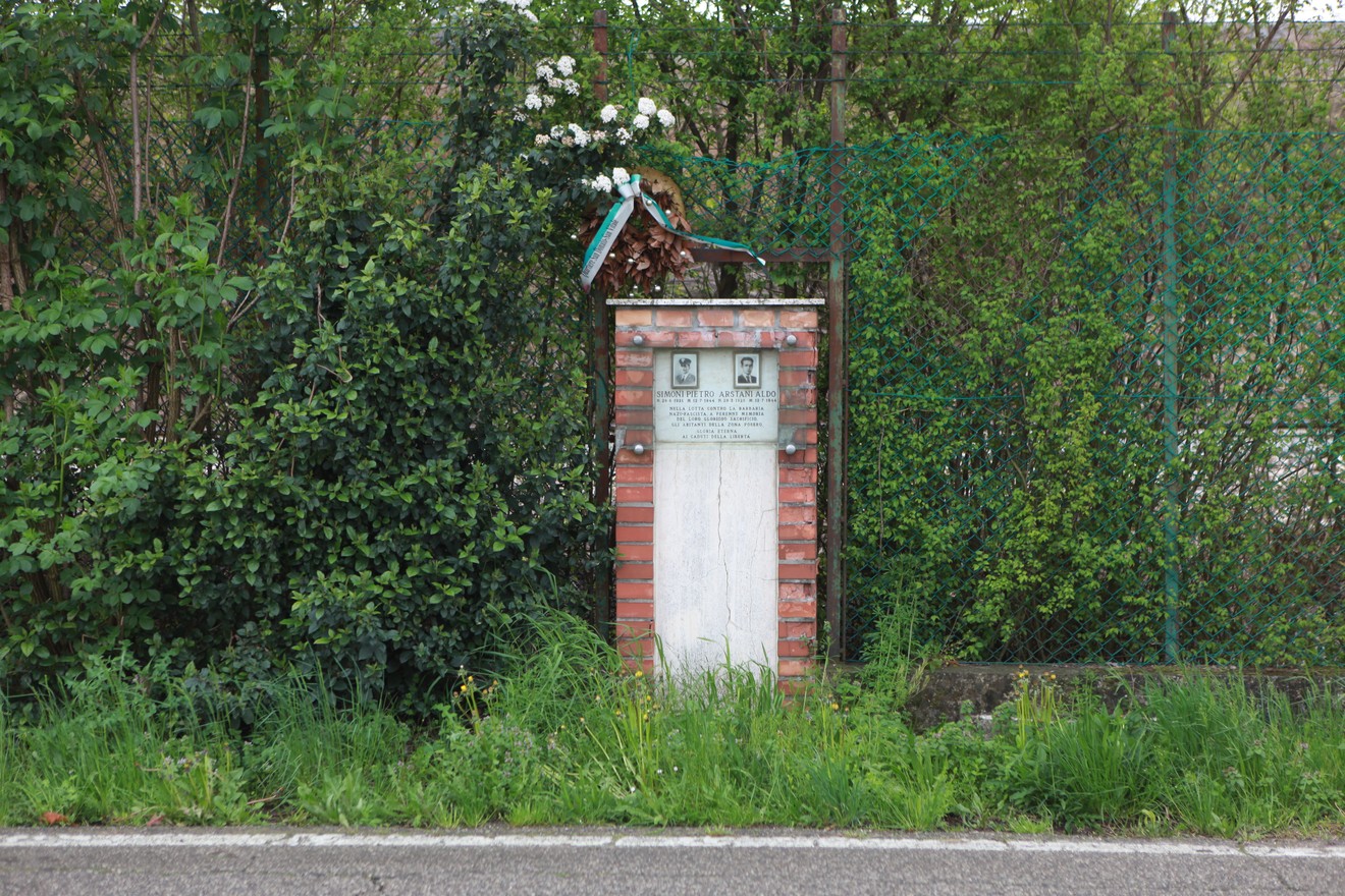 Cippo commemorativo circondato da vegetazione, con dettagli floreali e una parete in mattoni.
