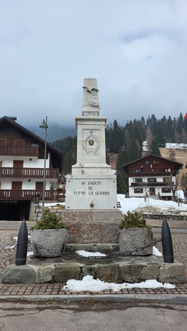 Monumento ai Caduti di Sappada, con base in pietra e statue, circondato da paesaggio montano e architetture in legno.