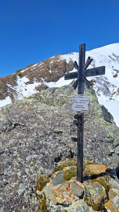 Croce metallica su una roccia coperta di muschio, con sfondo innevato e cielo blu chiaro.