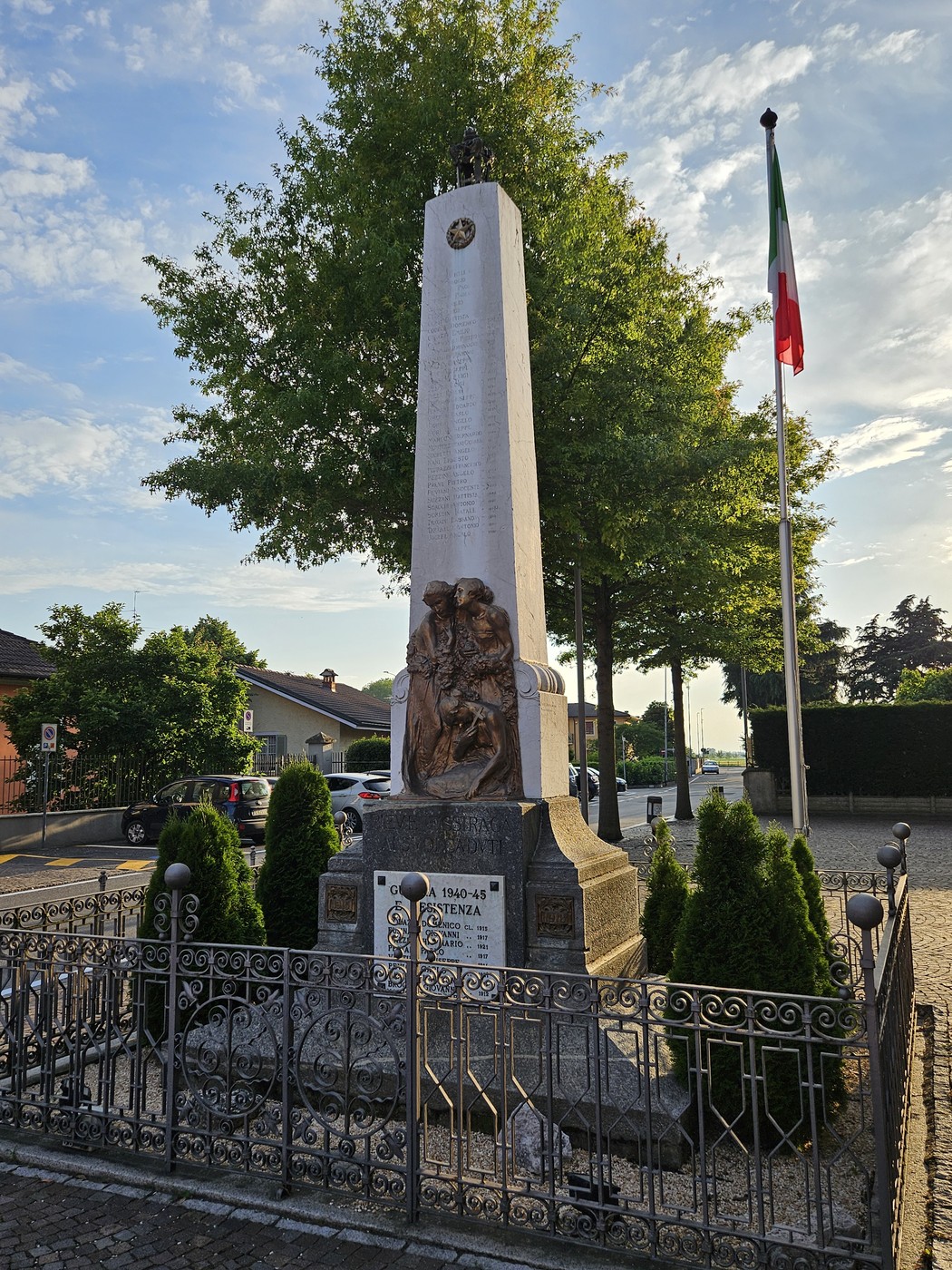 Monumento ai Caduti di Pieve Fissiraga, con obelisco e sculture commemorative, circondato da verde e bandiera italiana.