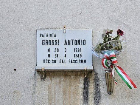 Lapide commemorativa dedicata a Antonio Grossi, patriota ucciso dal fascismo, con fiori e nastri tricolori.