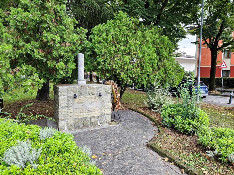 Cippo in pietra con colonna e iscrizione, in un'aiuola alberata con sentiero pavimentato.