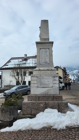 Monumento ai Caduti di Sappada, immerso in un paesaggio innevato. Sullo sfondo, case alpine e montagne.