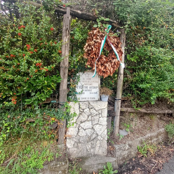 Cippo in pietra con lapide incisa, tra la vegetazione, sormontato da una corona con nastro tricolore.