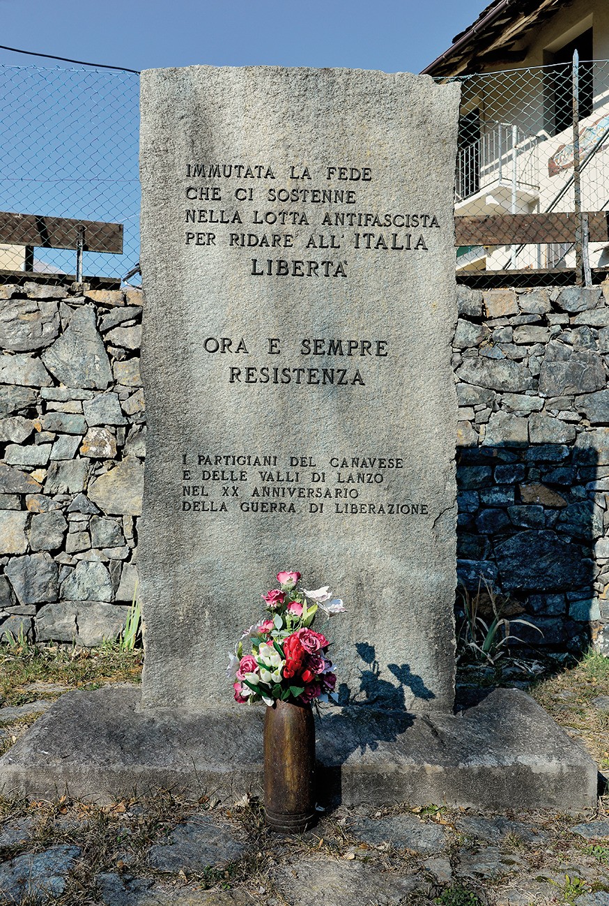 Cippo in pietra con iscrizione, fiori deposti in un vaso alla base, su sfondo di muro in pietra e cancellata.