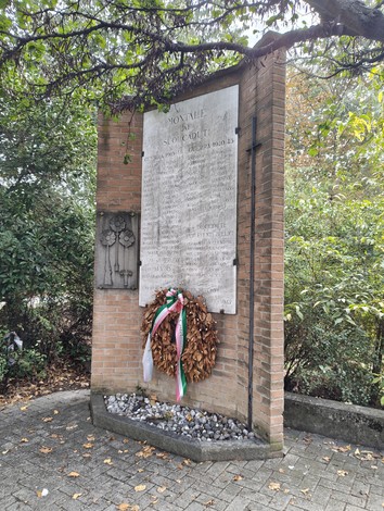 Monumento in mattoni con lapide incisa, decorazione in ferro battuto e corona con nastro tricolore, tra gli alberi.