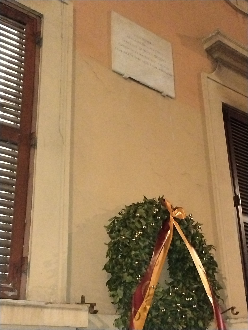 Lapide commemorativa con corona di alloro e nastri, affissa a un muro in un contesto urbano.