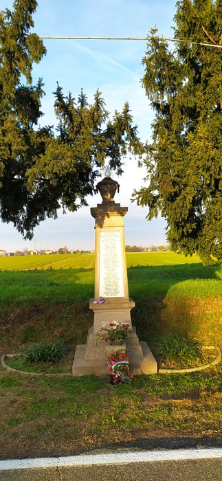 Monumento ai caduti dell'Amola immerso in un paesaggio rurale, con alberi e un campo sullo sfondo.