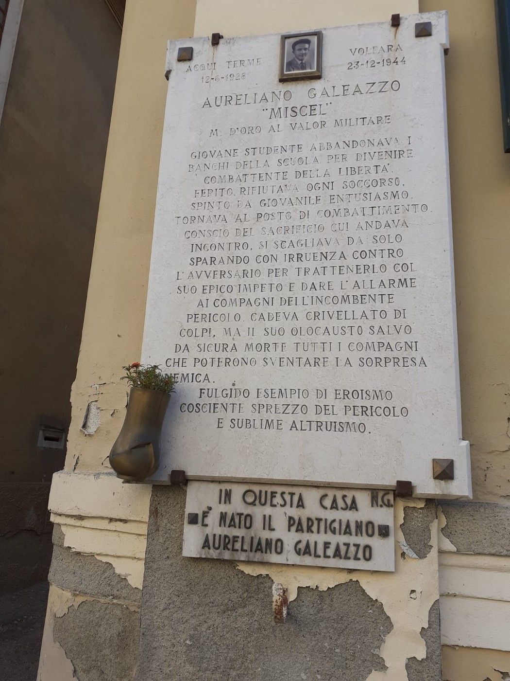 Lapide commemorativa di Aureliano Galeazzo, partigiano, con inscrizioni storiche e un omaggio floreale.