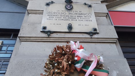 Lapide commemorativa di Leopoldo Montanucci con corona di alloro e nastro tricolore, situata in un contesto urbano.