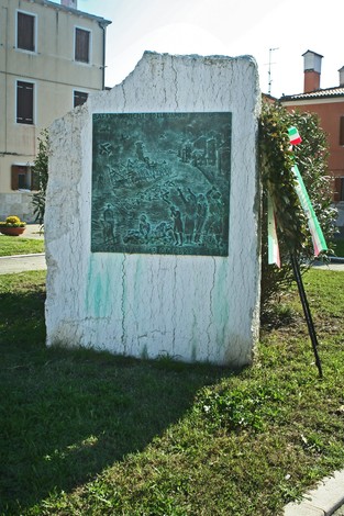 Monumento per l'affondamento del “Giudecca” con rilievi bronzei, situato in un contesto verde e urbano.