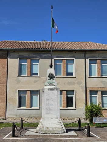 Monumento ai Caduti di Pezzoli, con bandiera italiana, accanto a un edificio storico e un'area verde.