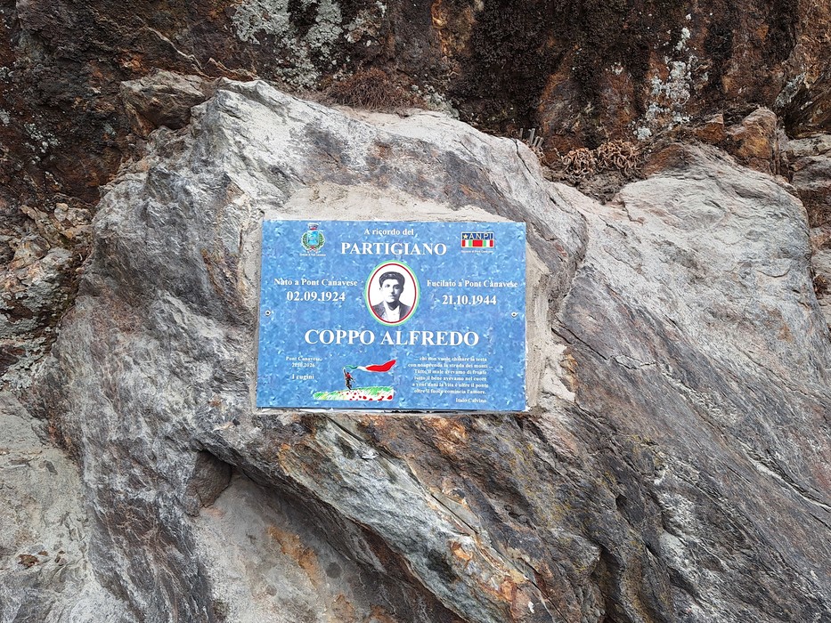 Targa blu con ritratto e iscrizione dedicata al partigiano Coppo, fissata su una roccia.