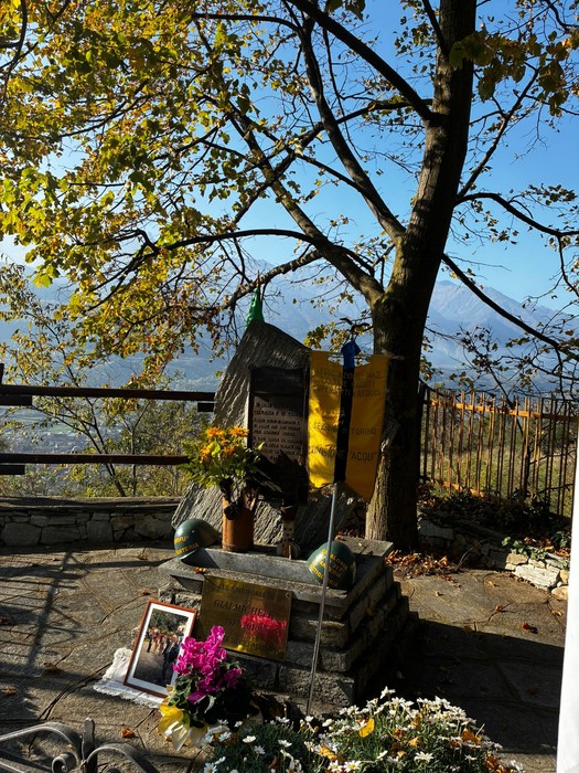 Monumento in pietra con iscrizione e drappo giallo, circondato da alberi, con fiori e foto deposti alla base.