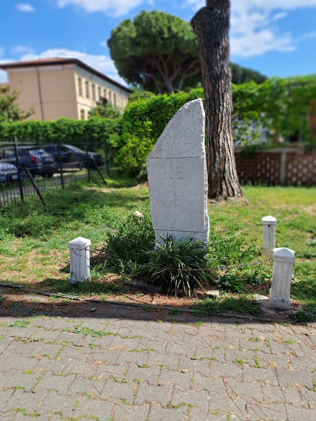 Cippo commemorativo del bombardamento di Avenza, circondato da vegetazione e delimitato da piccoli pilastri bianchi.