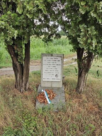 Cippo commemorativo avvolto da alberi, con decorazioni floreali e un paesaggio verde sullo sfondo.