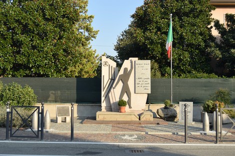 Monumento in pietra con sagoma di uomo a braccia alzate e iscrizione, in un'aiuola su strada con bandiera italiana.