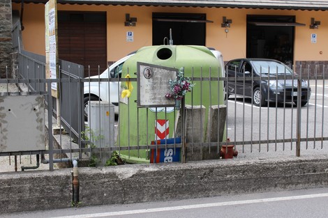 Lapide commemorativa su un supporto vicino a un contenitore per rifiuti, circondata da fiori e recinzione.