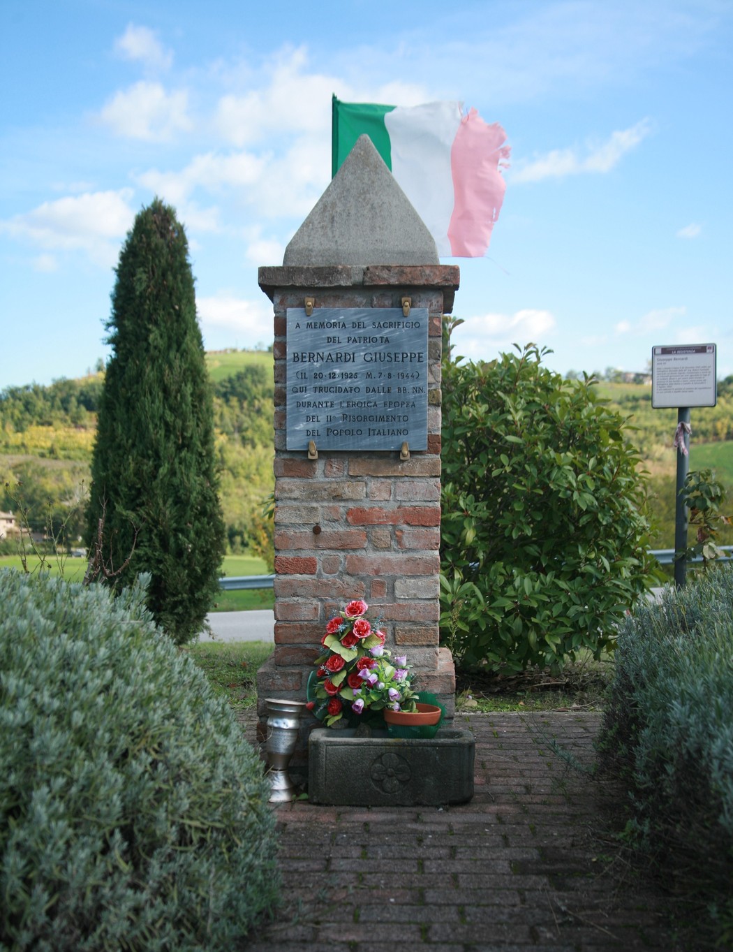 Cippo commemorativo con fiori e bandiera italiana in un paesaggio collinare.