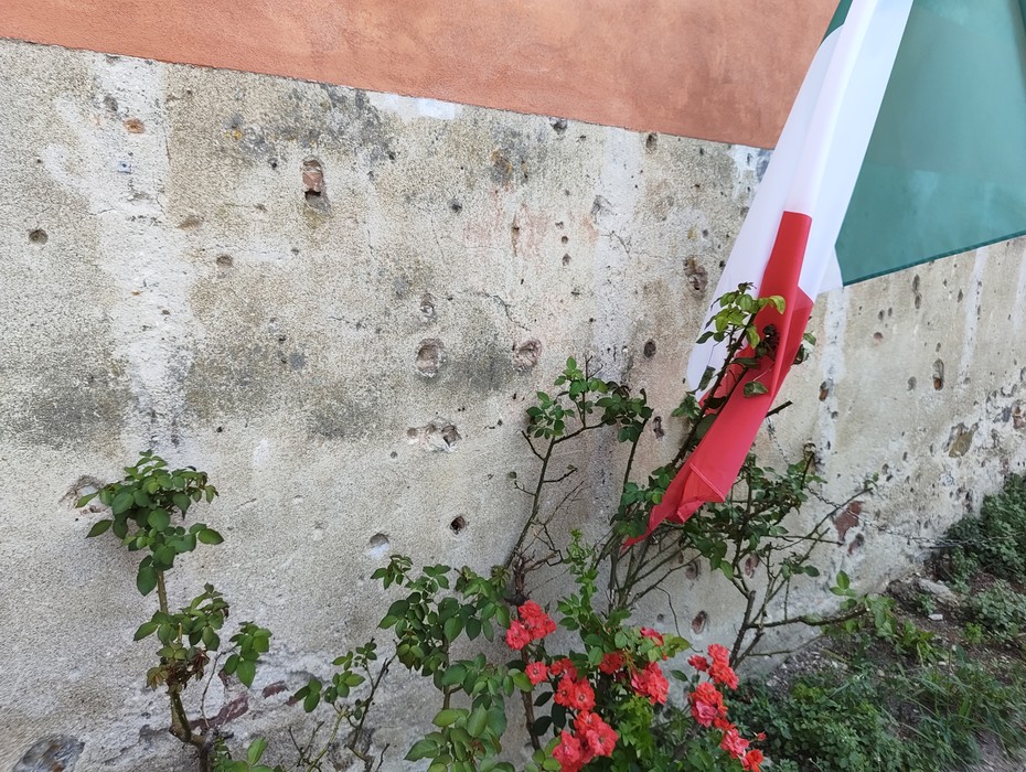 Muro storico con segni di proiettile, bandiera italiana e fiori rossi in primo piano.