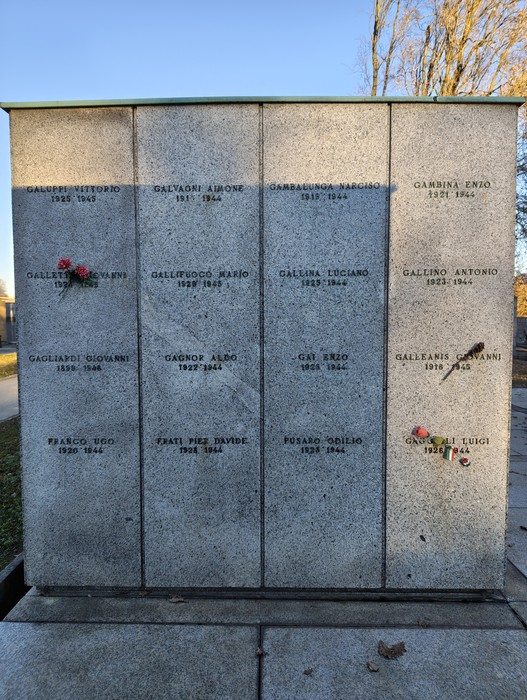 Monumento commemorativo con nomi incisi su lastre di pietra, adornato da fiori e bandiere.