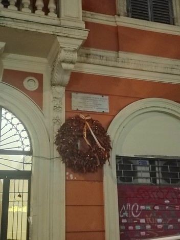 Lapide commemorativa con corona di alloro, affissa su un edificio di colore arancione.