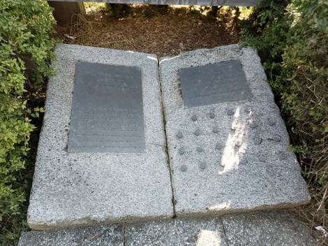 Monumento a D'Acquisto, due lastre di pietra commemorate con testi e forme, circondato da vegetazione.