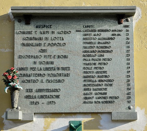 Lapide in pietra su parete, con iscrizione e doppio elenco di nomi; sotto, un vaso con fiori.