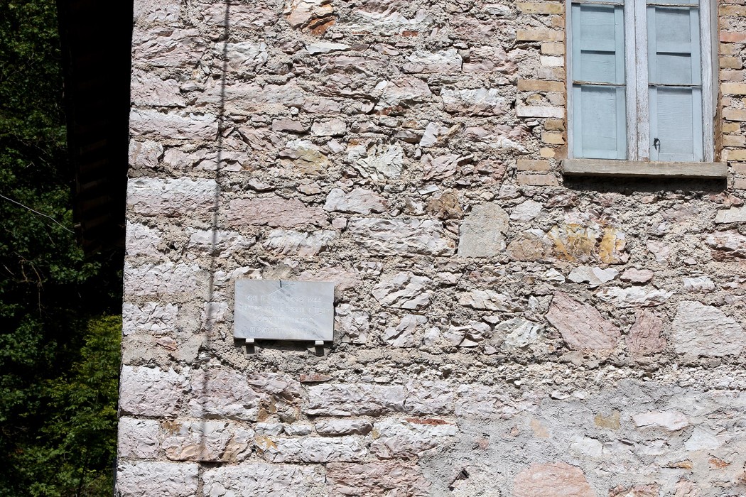 Lapide commemorativa su un muro di pietra, con finestre in legno sullo sfondo.