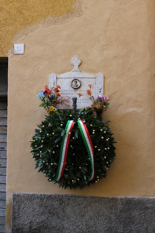 Lapide commemorativa adornata con un omaggio floreale, situata su una parete gialla.