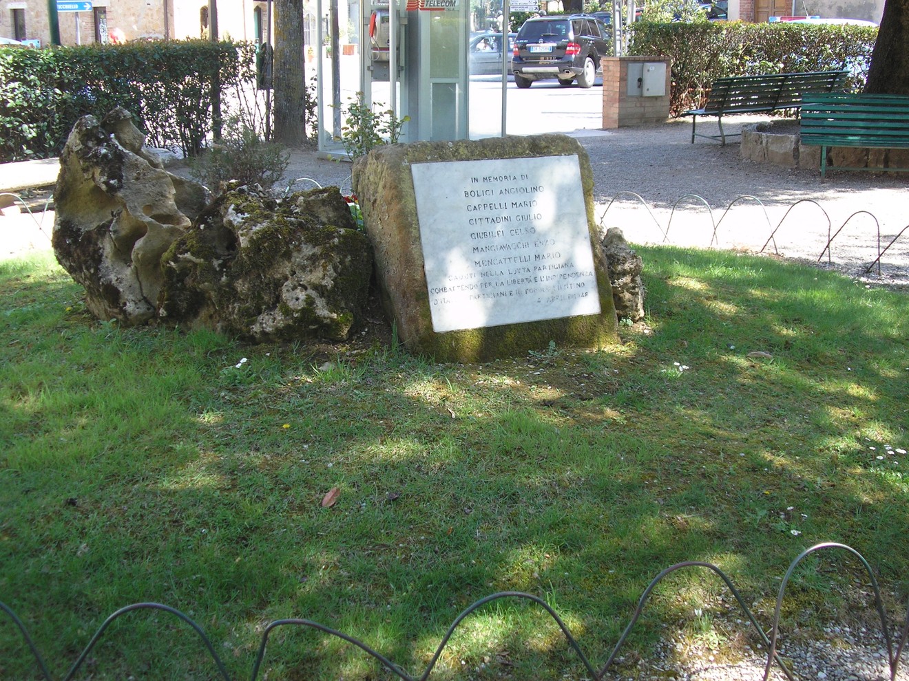 Cippo commemorativo ai partigiani, situato in un giardino pubblico, circondato da erba e recinzione decorativa.