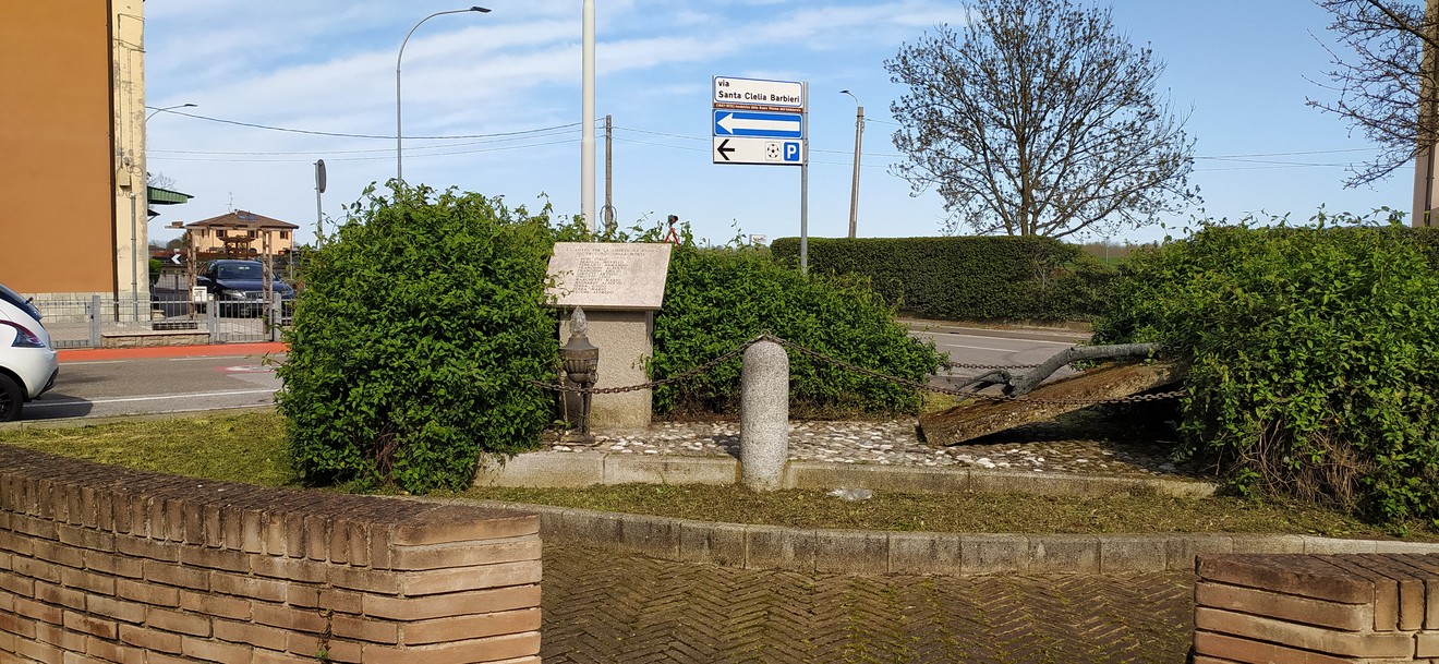 Stele commemorativa ai Caduti di Le Budrie, circondata da vegetazione, con segnaletica stradale sullo sfondo.