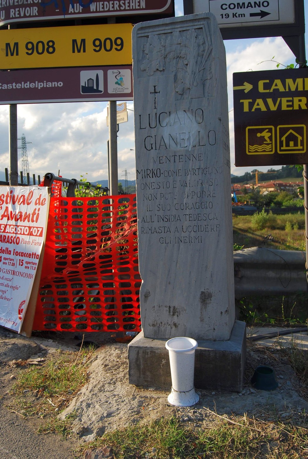 Cippo commemorativo dedicato a Gianello, con scritte e segnaletica stradale sullo sfondo.