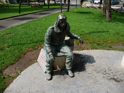 Monumento a Rampinelli, scultura che raffigura un uomo seduto con occhiali e giacca, immerso in un contesto verde.