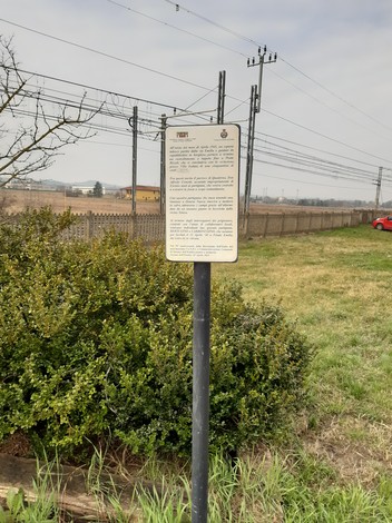 Targa informativa su un rastrellamento, situata in un'area aperta con vegetazione e linee elettriche sullo sfondo.