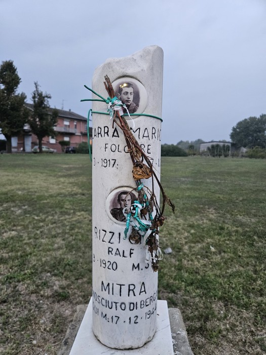 Cippo in pietra con iscrizione e foto dei caduti, ornato da fiori e nastri, su prato all'aperto.