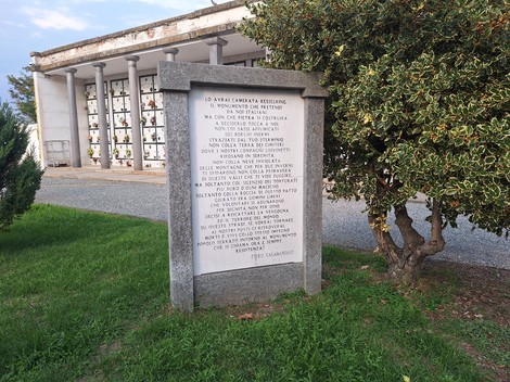 Lapide in pietra con iscrizione, posta su basamento erboso davanti a un portico, accanto a un albero.