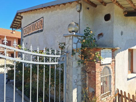 Lapide commemorativa su un muro di una casa, con vegetazione circostante e cancellata in metallo.