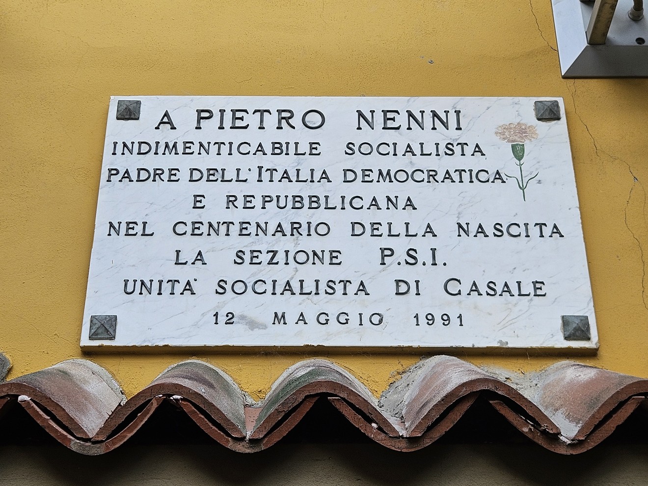 Lapide bianca con iscrizione dedicata a Pietro Nenni, fissata su una parete gialla sopra un tetto in coppi.