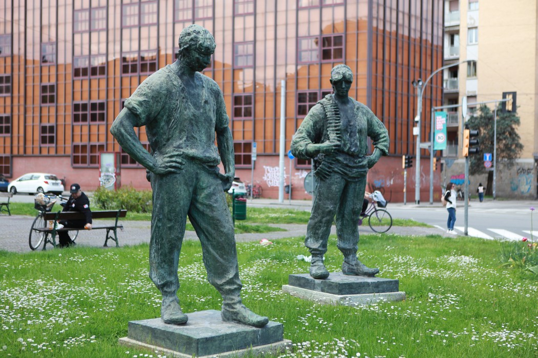 Due statue di partigiani in un parco, simboleggiano la lotta e il coraggio della Resistenza. Sullo sfondo, un edificio moderno.