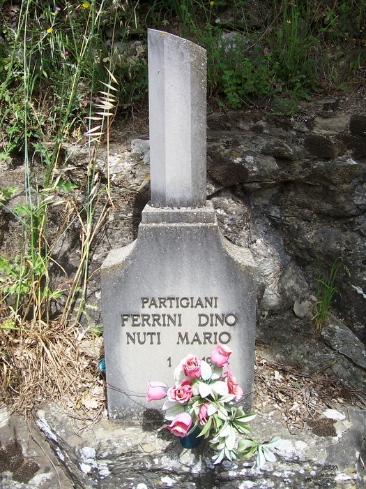 Cippo commemorativo dedicato ai partigiani Ferrini e Nuti, con fiori, in un ambiente naturale.